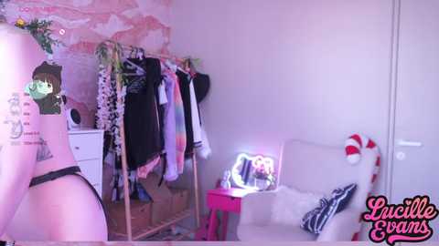 Media: Video of a young woman's bedroom with pastel walls, pink floral wallpaper, and a white bed. A pink stool holds a pink and white striped cushion. Clothes hang on a wooden rack.