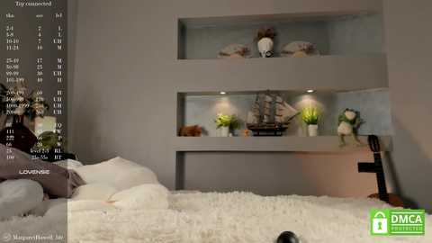 Media: Video of a modern bedroom with a bed covered in white bedding, a cozy white rug, and a gray wall featuring built-in shelves with nautical decor and potted plants.