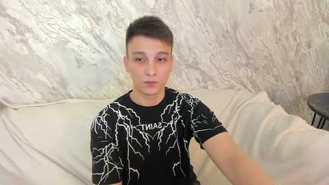 Media: Video of a young man with short dark hair, light skin, and a slim build, wearing a black T-shirt with lightning bolt designs. He sits on a beige couch against a textured, light gray wall, holding a phone for a selfie.