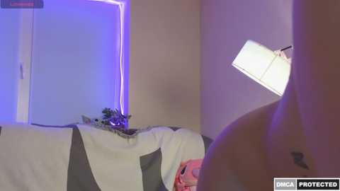 Media: A video of a nude woman with a small tattoo on her lower back, lying on a bed with a gray and white striped blanket, illuminated by blue light.