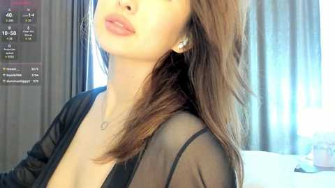Media: Video of a light-skinned woman with long brown hair, wearing a sheer black top, standing indoors with gray curtains and a bed visible in the background.