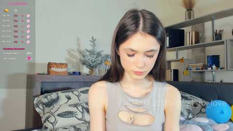 Media: Video of a young woman with long brown hair in a gray tank top, sitting on a bed with a floral duvet, surrounded by a cluttered room with a calendar, books, and a blue stress ball.