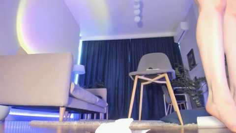 Media: Video of a modern, minimalist living room with a beige sofa, dark curtains, a gray chair, and a green potted plant. A rainbow arch appears in the upper left corner.