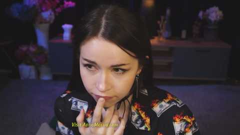 Media: Video of a young woman with long dark hair, wearing a black shirt adorned with dollar bills, in a dimly lit room with floral arrangements, holding her fingers to her lips.