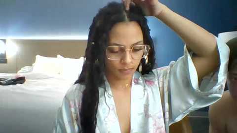 Media: Video of a fair-skinned woman with long black hair, wearing large glasses and a light robe, adjusting her hair in a modern bedroom with a bed and blue wall.