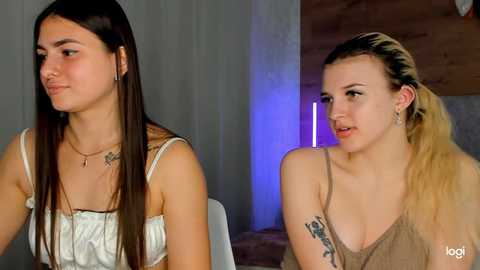 Media: Video of two young women with long hair, one in a white top and the other in a beige tank, both with tattoos and sitting indoors.