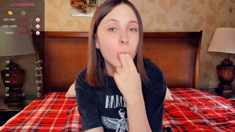 Media: Video of a young woman with fair skin and straight brown hair, lying on a bed with a red plaid blanket. She's wearing a black t-shirt and is seductively biting her finger. The background features a wooden headboard and a beige, floral-patterned wallpaper.