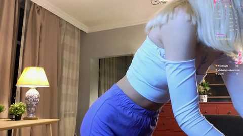 Media: Video of a blonde woman wearing a white off-the-shoulder crop top and blue pants, bending over in a room with beige curtains, a red dresser, and a lamp.