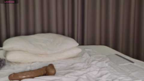 Media: A video of a messy bed with white sheets, a white pillow, and a brown dildo on the left. Dark brown curtains in the background.