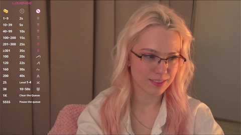 Media: Video of a smiling, fair-skinned woman with long, wavy platinum blonde hair and glasses, wearing a white blouse and pink cardigan, sitting indoors with a wooden panel background.
