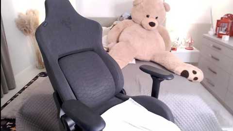Media: Video of a beige teddy bear lounging on a black gaming chair in a minimalist, light-colored bedroom with a white dresser and lamp.