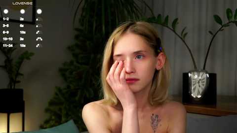 Media: Video of a young, pale-skinned woman with shoulder-length blonde hair, wearing a blue hair clip, sitting indoors, topless, with a pensive expression. Background includes green plants, a black lamp, and a wooden shelf.