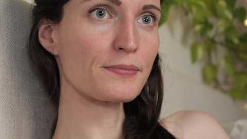 Media: A video of a young woman with fair skin, blue eyes, and long brown hair, wearing a light gray top. She gazes to the side, with blurred greenery in the background.