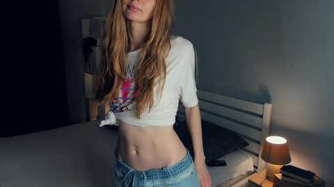 Media: Video of a slender, fair-skinned woman with long, straight, auburn hair, wearing a cropped white T-shirt and low-rise jeans, standing in a dimly lit bedroom with a lamp, bed, and wall art.