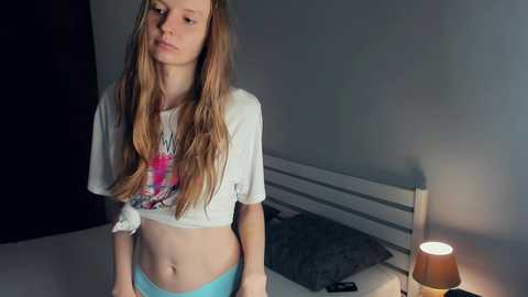 Media: Video of a young Caucasian girl with long, wavy, light brown hair, wearing a white cropped graphic T-shirt and blue shorts. She stands near a bed with a grey pillow and a bedside lamp casting a warm glow in a dimly lit room.