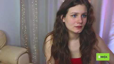 Media: A video shows a young woman with long, wavy brown hair, wearing a red top, seated indoors. She has a neutral expression. Background features a beige leather couch and semi-transparent curtains with small lights.