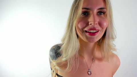 Media: Video of a smiling, fair-skinned woman with blonde hair and a tattoo on her shoulder. She wears a strapless dress and a heart pendant necklace.