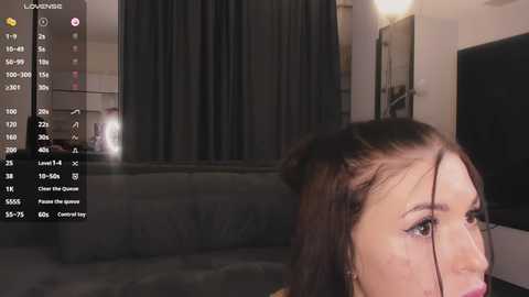 Media: A video of a woman with long, dark hair, wearing makeup, captured from a low-angle, showing a dimly lit room with a large TV screen and dark curtains in the background.