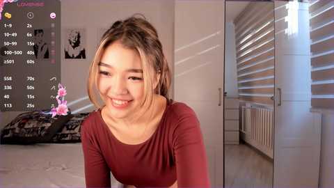 Media: Video of a young Asian woman with light skin and long, blonde hair smiling in a modern, brightly lit bedroom with white walls, a bed, and blinds.