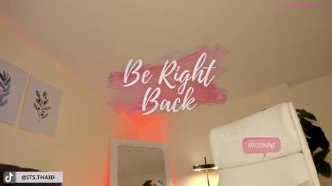 Media: Video of a cozy, well-lit room with a white fridge, a red-tinted lamp, and a pink \"Be Right Back\" sign on the wall, alongside two white framed floral prints.