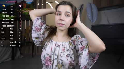 Media: Video of a young Caucasian woman with fair skin, brown hair, and a floral blouse, standing in a dimly lit room. She has a relaxed, casual pose.