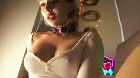 Media: Video of a blonde woman with large breasts, wearing a low-cut beige top, black choker, and a pink neon sign in the background.