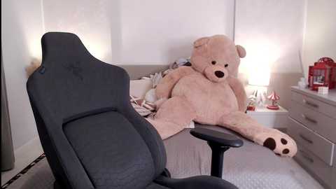 Media: A video of a beige teddy bear lounging on a grey gaming chair in a minimalist bedroom with a white bed, dresser, and lamp.