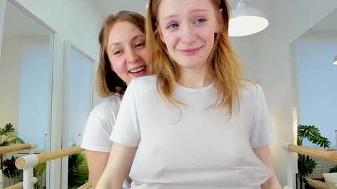 Media: Video of two smiling young women with light skin, long hair, and white t-shirts, standing in a modern, bright room with white walls, plants, and a wooden railing.