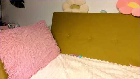 Media: Video of a child's bed with a mustard-yellow headboard, a pink, fringed pillow, and a white, textured blanket. Background features a cream-colored teddy bear and a large, colorful flower.