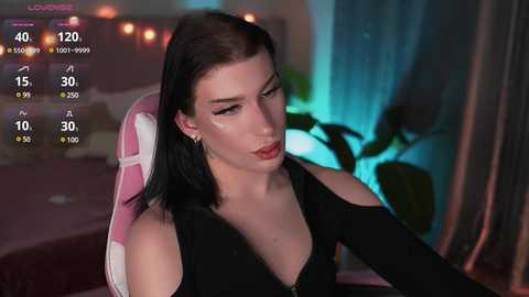 Media: Video of a young woman with pale skin, black hair, and red lipstick, wearing a black top, sitting in a pink chair, in a dimly lit room with a blue-green background.