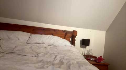 Media: Video of a simple, dimly-lit attic bedroom with a wooden headboard bed, white bedding, a black lamp, and a red item on the wooden nightstand against a white wall.