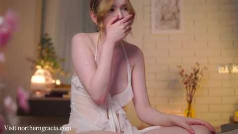 A young woman with fair skin and blonde hair, wearing a sheer white dress, sits on a bed, covering her mouth in a shocked expression. The background features a white brick wall and a vase of dried flowers.