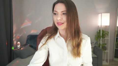 Media: Video of a fair-skinned woman with long brown hair, wearing a white button-down shirt, smiling, seated indoors in a modern, softly lit room with a tall floor lamp.