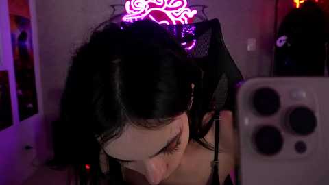 Media: Video of a young woman with long black hair, wearing headphones, taking a selfie in a dimly lit room with a neon pink sign and a large iPhone camera.