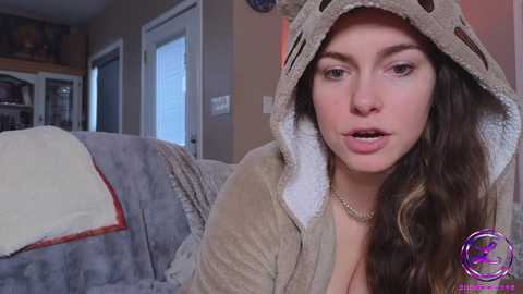 Media: Video of a young woman with long brown hair, wearing a beige, plush, hooded animal costume, sitting on a gray couch in a cozy, dimly lit living room.