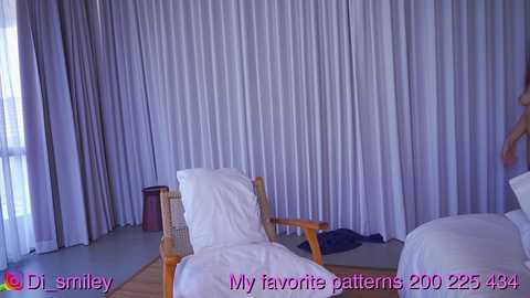 Media: A video of a minimalist bedroom with white curtains, a wooden chair, and a bed covered in white linens.