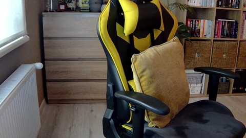 Media: Video of a yellow and black gaming chair with a beige cushion, positioned in a cozy, organized room with wooden furniture, bookshelves, and potted plants.