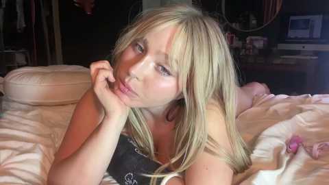 Media: Video of a fair-skinned, blonde woman with bangs, lying on a bed with white sheets, wearing a black strapless bra, in a dimly lit room with a computer and dark background.