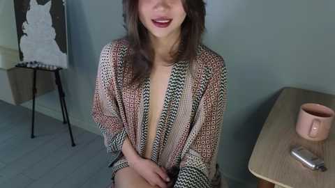 Media: Video of a young woman with light brown hair, smiling, wearing a sheer, patterned robe, seated in a modern, minimalist room with a wooden table, cup, and abstract art on the wall.
