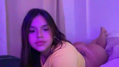 Media: Video of a young Asian woman with long black hair, wearing a light pink top, lying on her stomach on a bed, bathed in purple lighting, in a dimly-lit room.