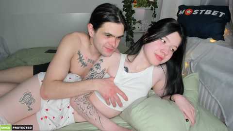 Media: Video of a shirtless man with tattoos and a woman with long black hair in white pajamas lying on a bed, smiling.
