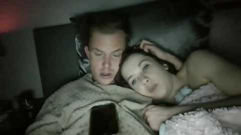 Media: Video of a man and woman, both Caucasian, sleeping on a bed. The man has short brown hair, and the woman has brown hair tied back. They are under a blanket, with a phone on the bed.