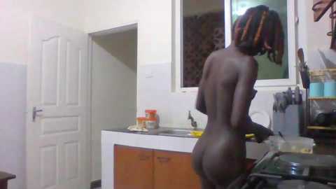 Media: Video of a nude woman with medium skin tone and braided hair, cooking at a kitchen counter, with a stove and white door in the background.