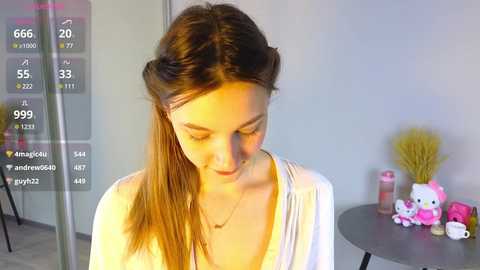 Media: A video of a young woman with long brown hair in a ponytail, wearing a white top, standing in a room with a grey table holding plush toys, and a digital overlay displaying social media metrics.