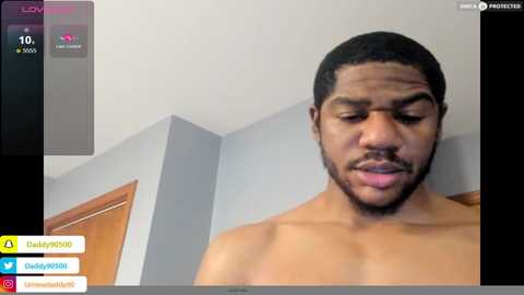 Media: Video of a shirtless Black man with a trimmed beard, looking serious, in a dimly lit room with grey walls and a wooden door.