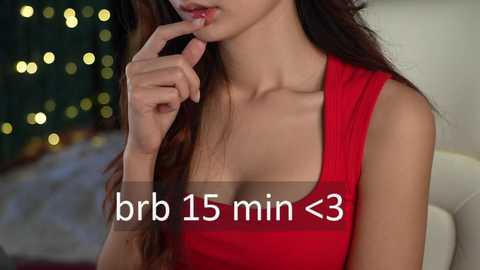 Media: Video of an Asian woman with long brown hair, wearing a red sleeveless top, holding her finger to her lips, with a blurred background featuring festive lights. Text reads \"brr 15 min <3.\