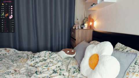 Media: Video of a cozy bedroom with a large, plush white sunflower pillow on a bed with floral sheets, a wooden nightstand, and a lit lamp beside a dark curtain.