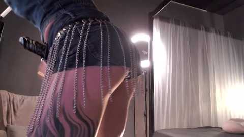 Media: Video of a woman wearing a tight, black-and-purple striped skirt with metallic chain details, posing provocatively, in a dimly lit room with a draped curtain and spotlight.