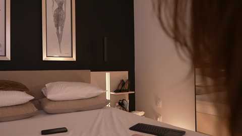 Video of a dimly-lit bedroom with a woman's long hair flowing, dark walls, framed drawings, beige bedding, pillows, bedside table with a phone, and a lamp.
