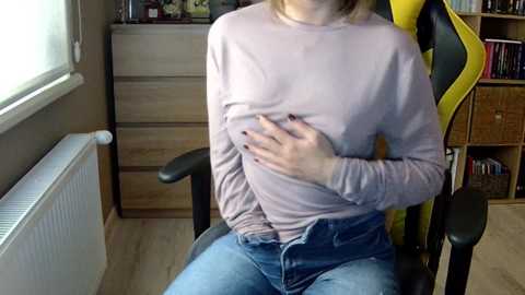 Media: Video of a woman in a purple sweater, blue jeans, seated in a black-and-yellow gaming chair, holding her chest, in a cluttered room with wooden furniture and bookshelves.